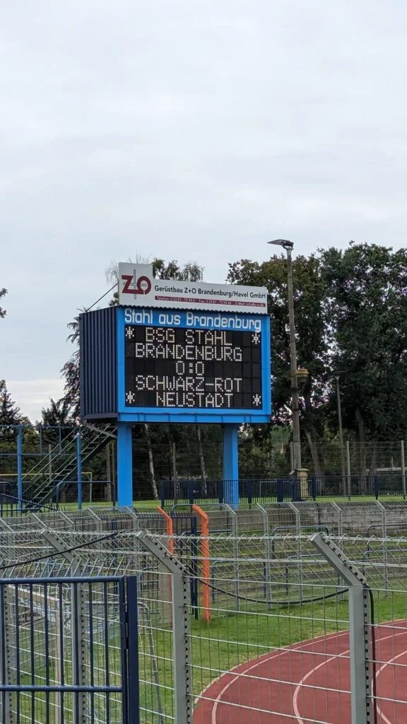 Anzeigetafel im Stadion am Quenz mit den Namen BSG Stahl Brandenburg und Schwarz-Rot Neustadt, davor die Laufbahn