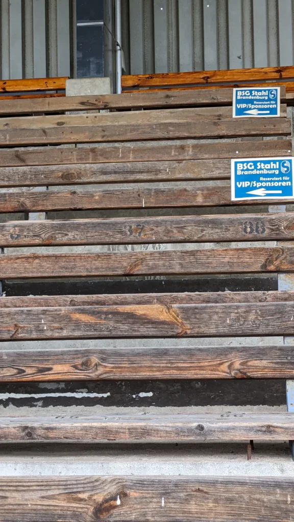 Verwitterte Holzbänke auf der Tribüne im Stadion am Quenz mit blau-weißen Platzschildern