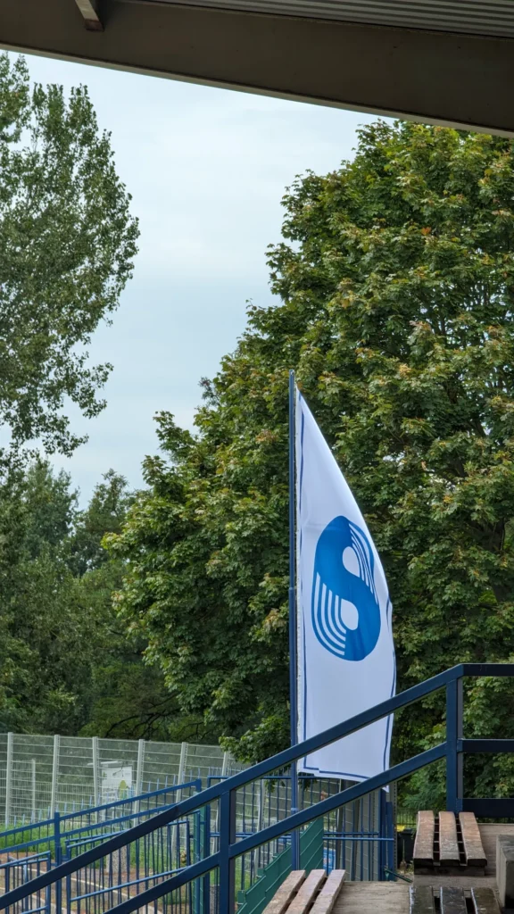 Weiße Fahne mit dem blauen Vereinszeichen von Stahl Brandenburg am Tribünenaufgang, dahinter Bäume