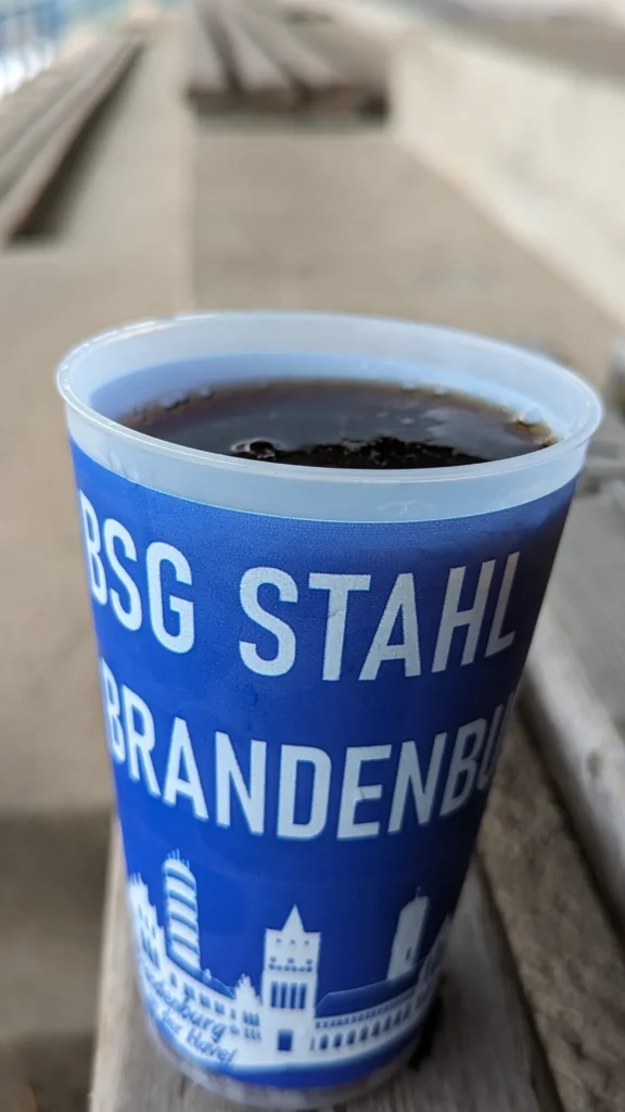 Blauer Becher mit der Aufschrift BSG Stahl Brandenburg auf einer Holzbank der Tribüne