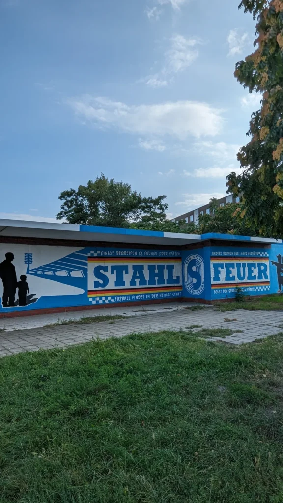 Blau-weißes Graffito Stahl Feuer mit Vereinswappen an einer Garagenwand, davor eine Wiese