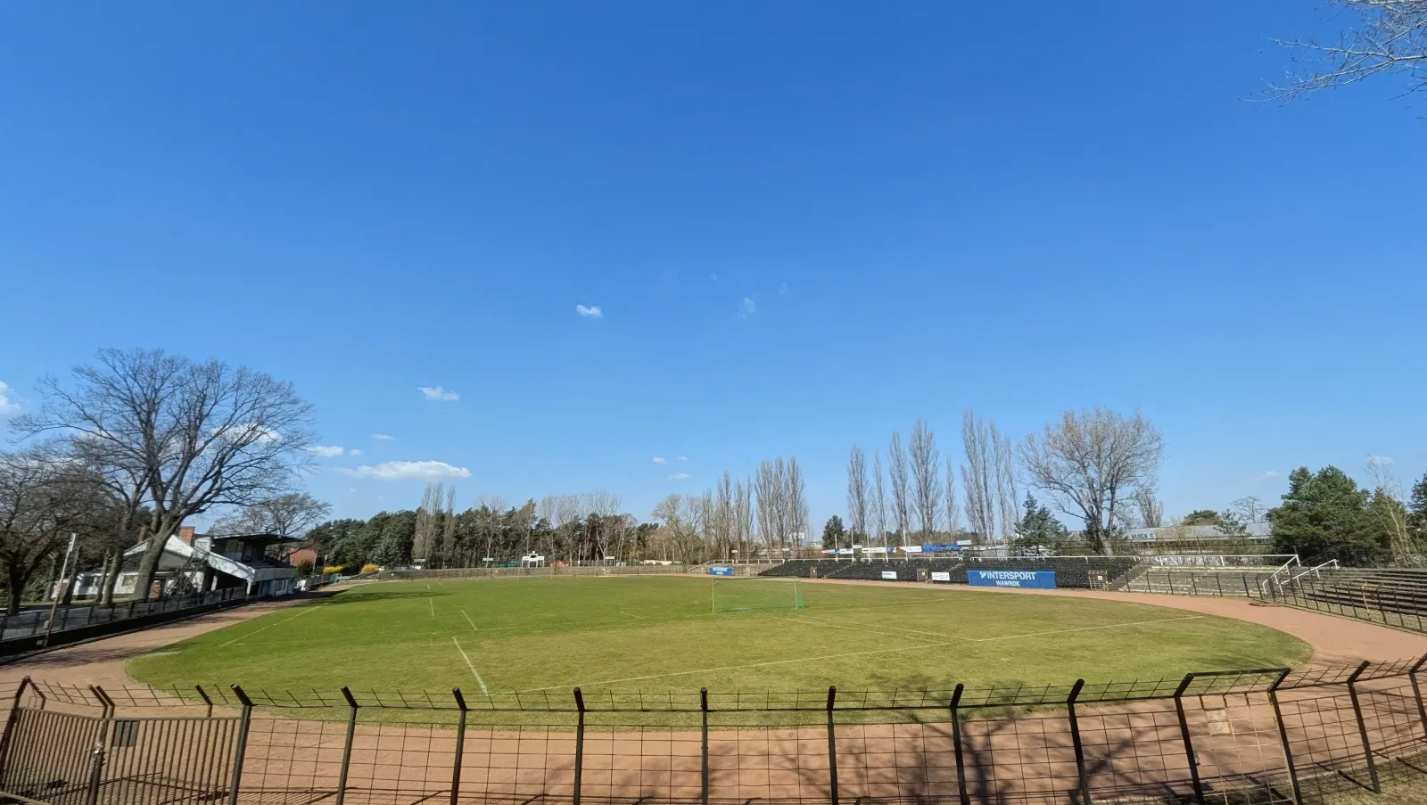 Blick über das Rasenspielfeld im Stadion der Hüttenwerker mit Laufbahn, Zaun und Kiefern unter blauem Himmel