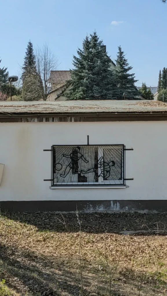 Weißes Nebengebäude am Stadion der Hüttenwerker mit einem schmiedeeisernen Fenstergitter, das Fußballer zeigt