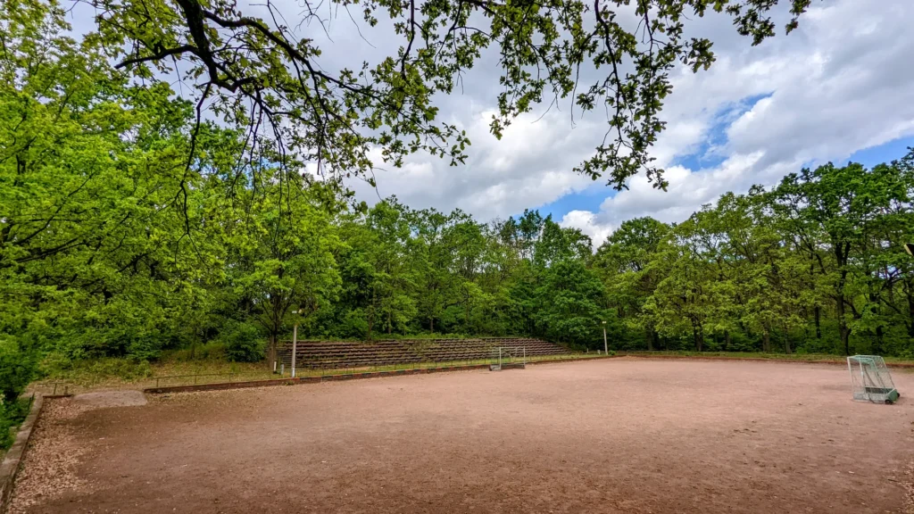 Ascheplatz mit Stufenreihen am Hang, Fußballtoren und Laubbäumen im Park Wuhlheide