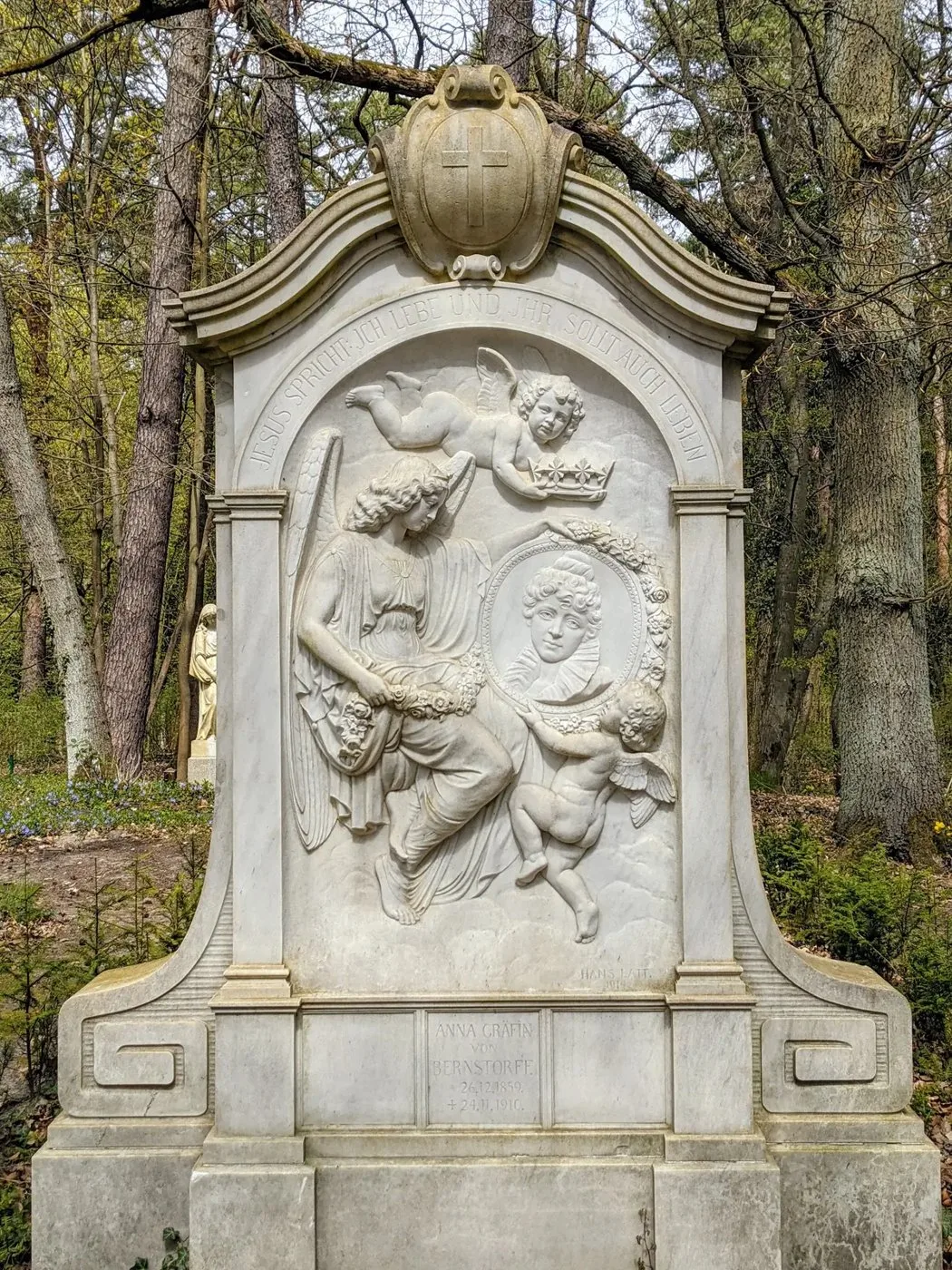 Weißes Grabmal mit Marmorrelief aus Figuren und Putten zwischen Bäumen