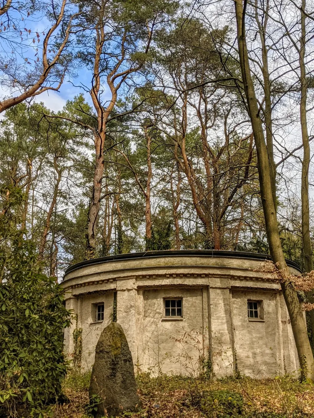 Runder Grabbau mit kleinen Fenstern im Wald, davor ein aufrecht stehender Stein
