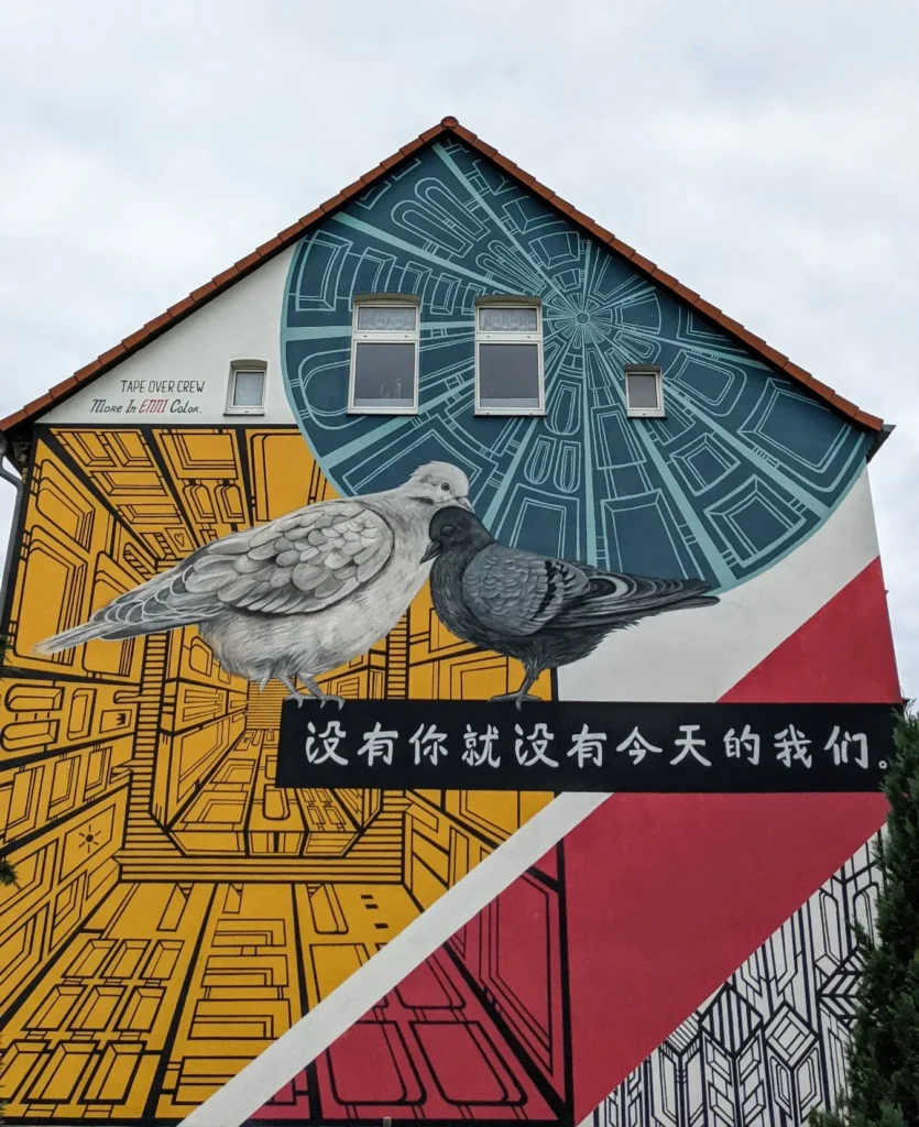 Wandbild Without you there is no us an der Berliner Straße in Lutherstadt Wittenberg: zwei Tauben vor gelben und roten Mustern, darunter ein Band mit chinesischen Schriftzeichen