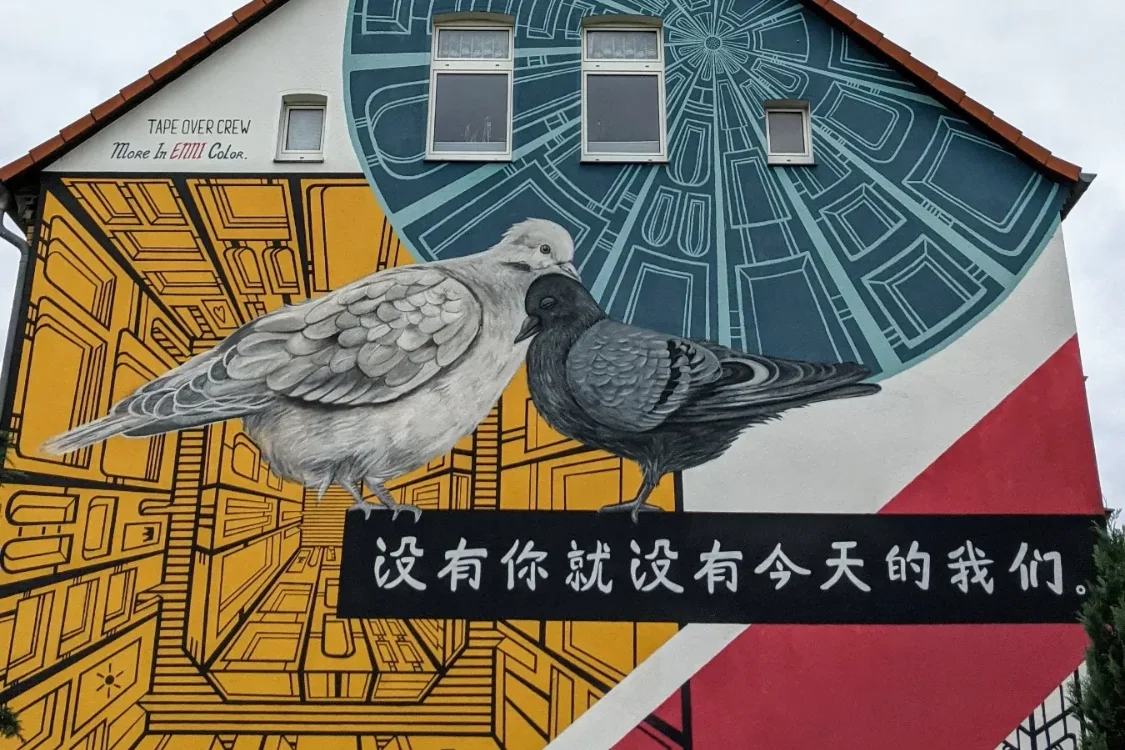 Wandbild „Without you there is no us“ an der Berliner Straße in Lutherstadt Wittenberg: zwei Tauben vor gelben und roten Mustern, darunter chinesische Schriftzeichen