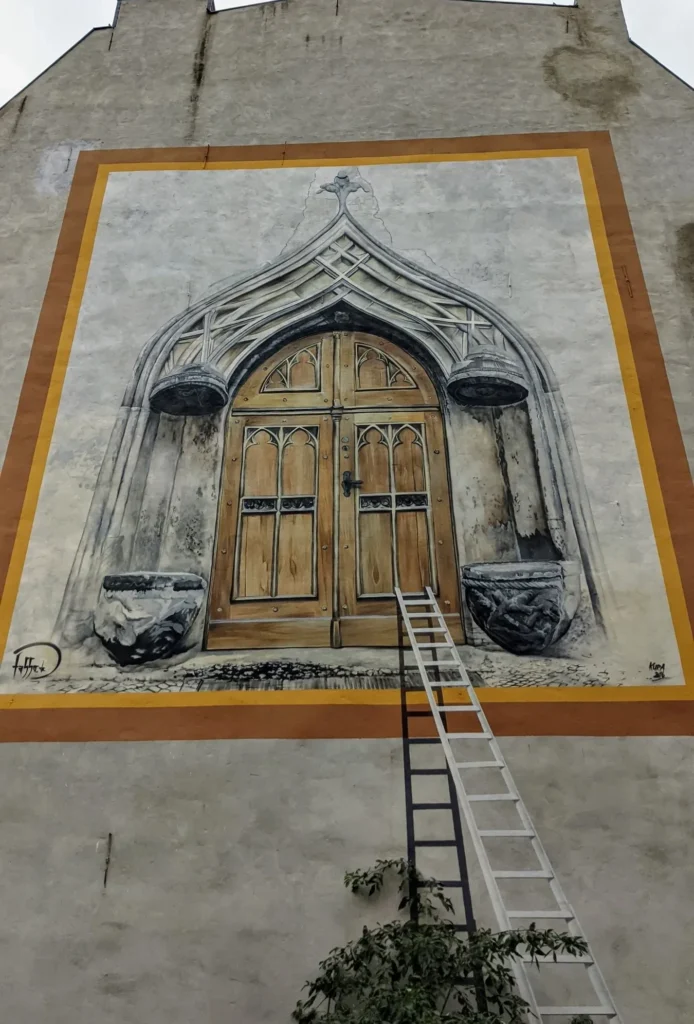 Wandbild von Tasso an der Coswiger Straße in Lutherstadt Wittenberg: gemaltes gotisches Holztor mit Steinrahmen und Leiter an einer Brandwand