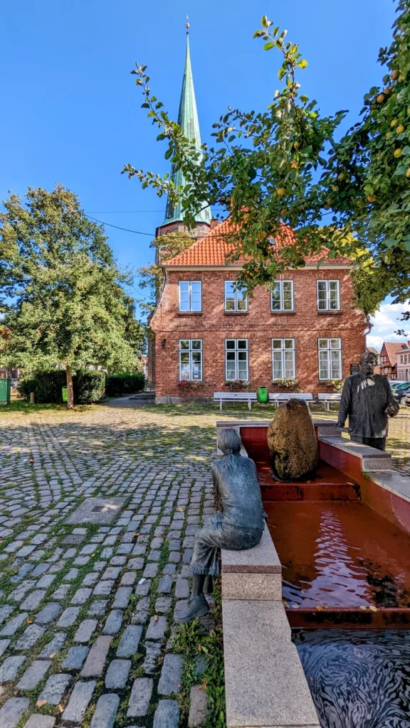 Brunnen mit Bronzefiguren am Alten Markt in Travemünde, dahinter ein Backsteinhaus