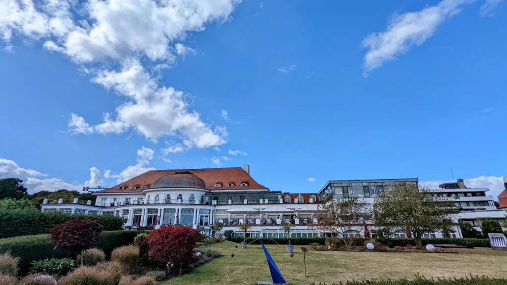 Blick über den Rasen des Kurgartens auf ein weißes Hotelgebäude mit rotem Dach in Travemünde
