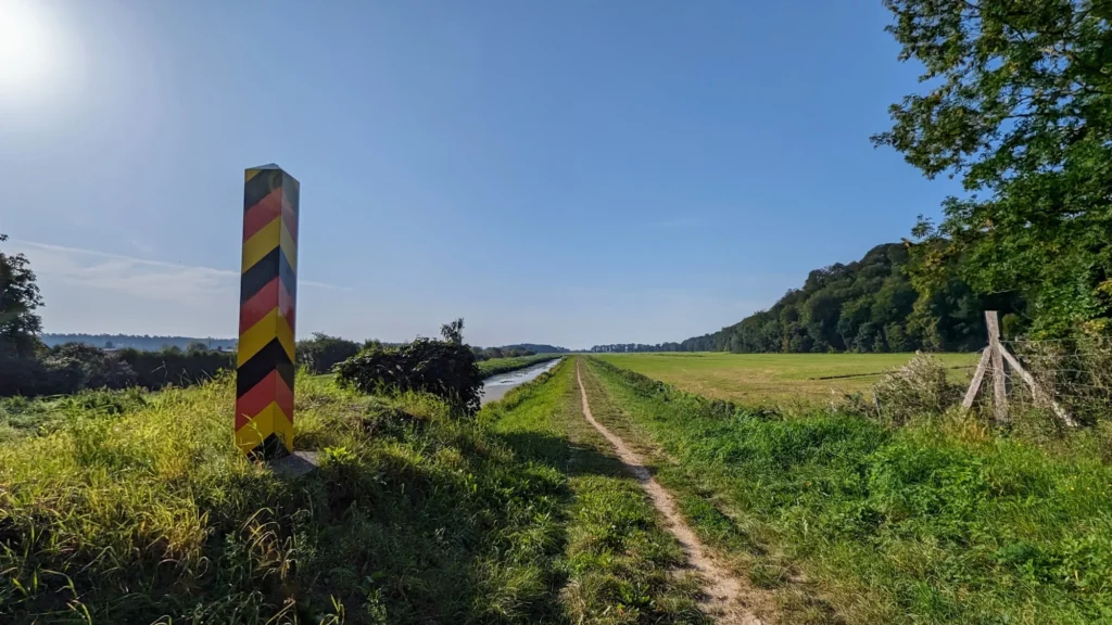 Schwarz-rot-goldener Grenzpfahl an einem Wiesenweg neben einem Graben, rechts ein bewaldeter Hang