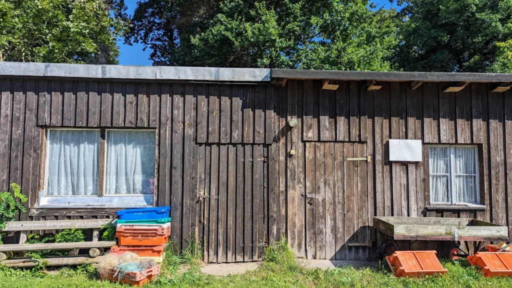 Dunkler Holzschuppen mit Gardinen, davor Fischkisten, Netze und ein Handwagen in Kölpinsee