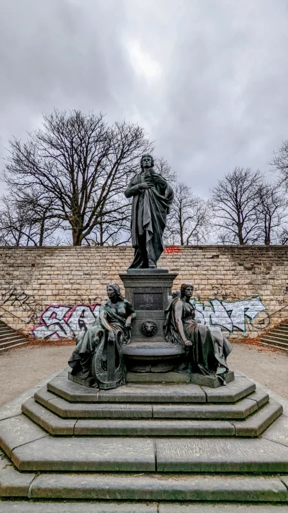 Bronzenes Schillerdenkmal mit sitzenden Frauenfiguren vor der Kalksteinmauer der Bastion im Schillerpark