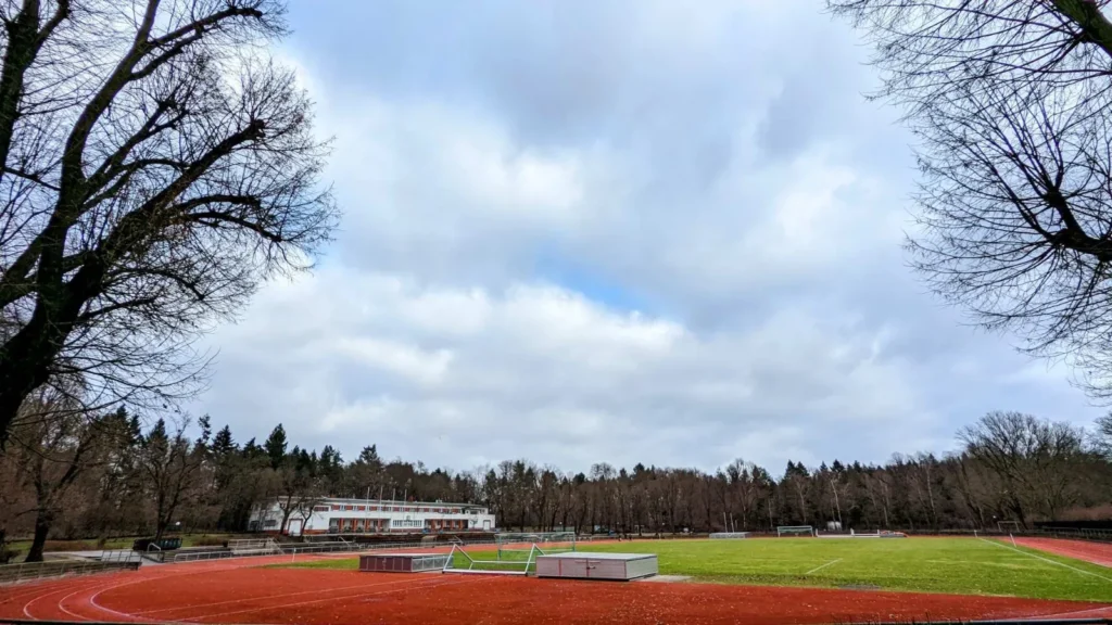 Laufbahn und Rasenplatz der Sportanlage im Volkspark Rehberge, dahinter ein Flachbau und Kiefernwald