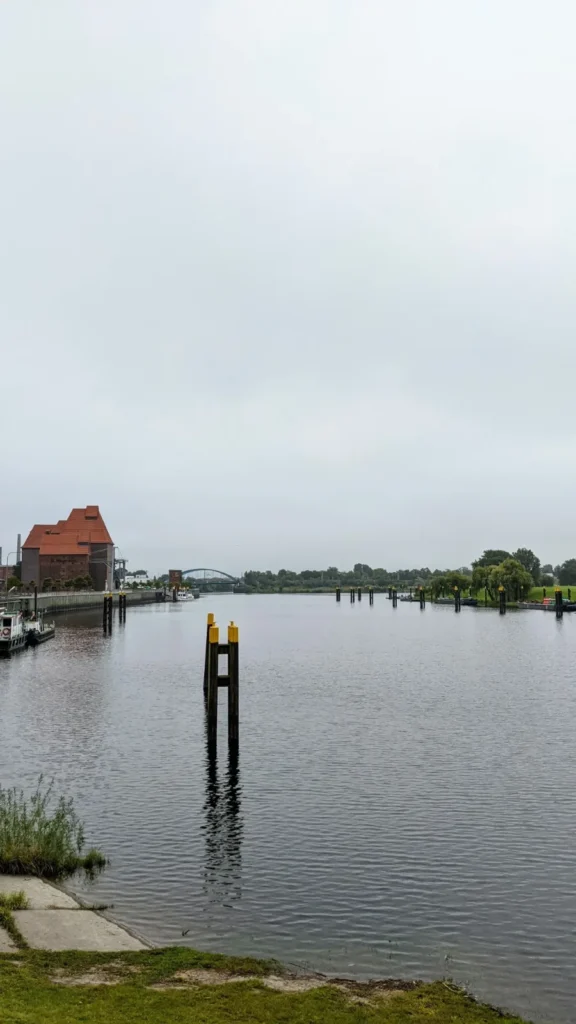 Hafenbecken in Wittenberge mit Pfahl im Wasser, im Hintergrund ein rotes Backsteingebäude und eine Brücke