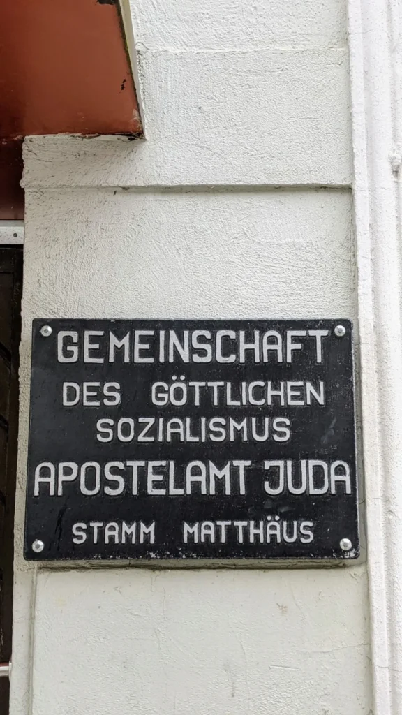 Schwarzes Schild an einer Hauswand in Wittenberge: Gemeinschaft des göttlichen Sozialismus, Apostelamt Juda, Stamm Matthäus