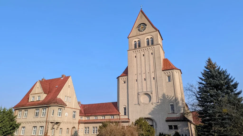 Hell verputzte Kirche St. Laurentius in Wriezen mit hohem Turm, Uhr und Satteldach, links das angebaute Pfarrhaus mit roten Dächern