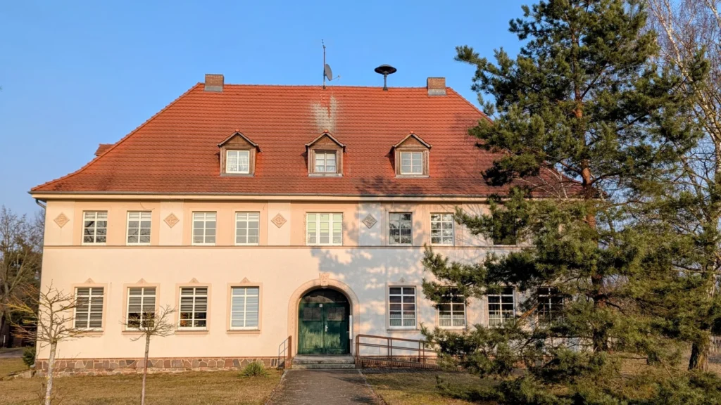 Zweigeschossiger, hell verputzter Bau mit hohem rotem Walmdach, drei Gauben und Rundbogenportal in Altranft, auf dem Dach eine Sirene