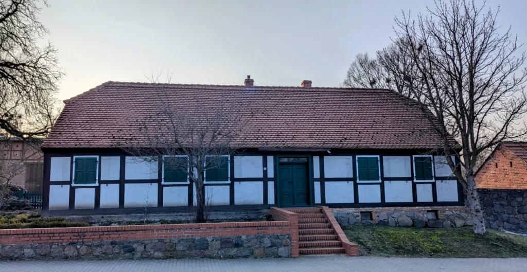 Eingeschossiges Fachwerkhaus mit weißen Gefachen, grünen Fensterläden, grüner Tür und rotem Ziegeldach in Altranft