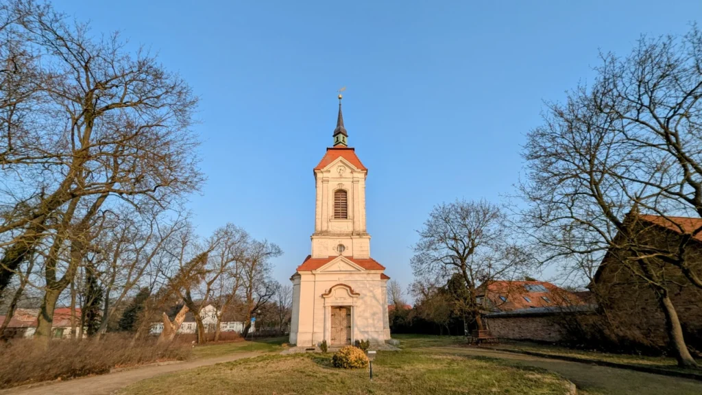 Dorfkirche Altranft mit schlankem, rosa-weißem Turm zwischen kahlen Bäumen